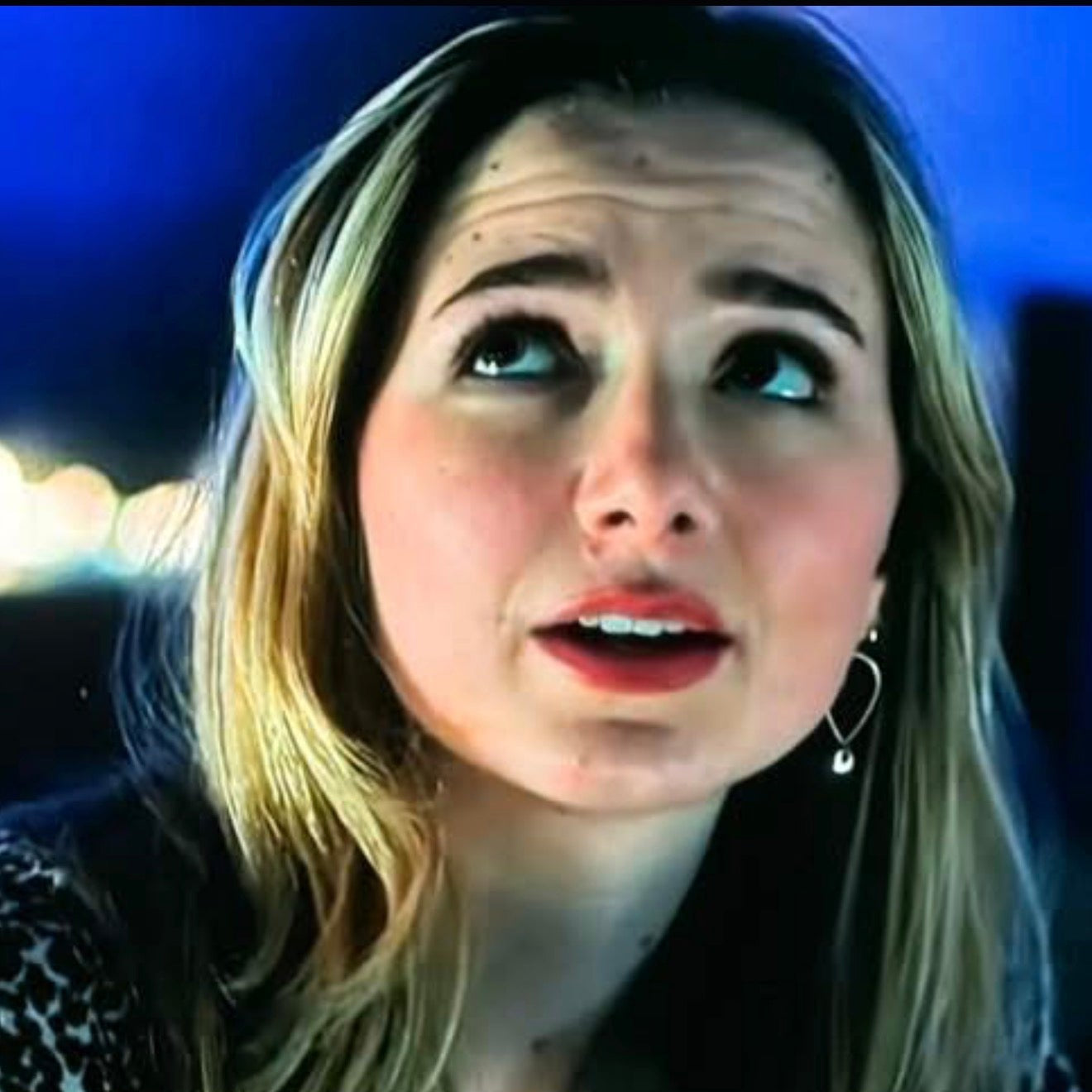 Woman with blonde hair and a leopard print top in a dark setting  wearing gold toned tear drop shaped earrings with silver toned bead dangling from the bottom.
