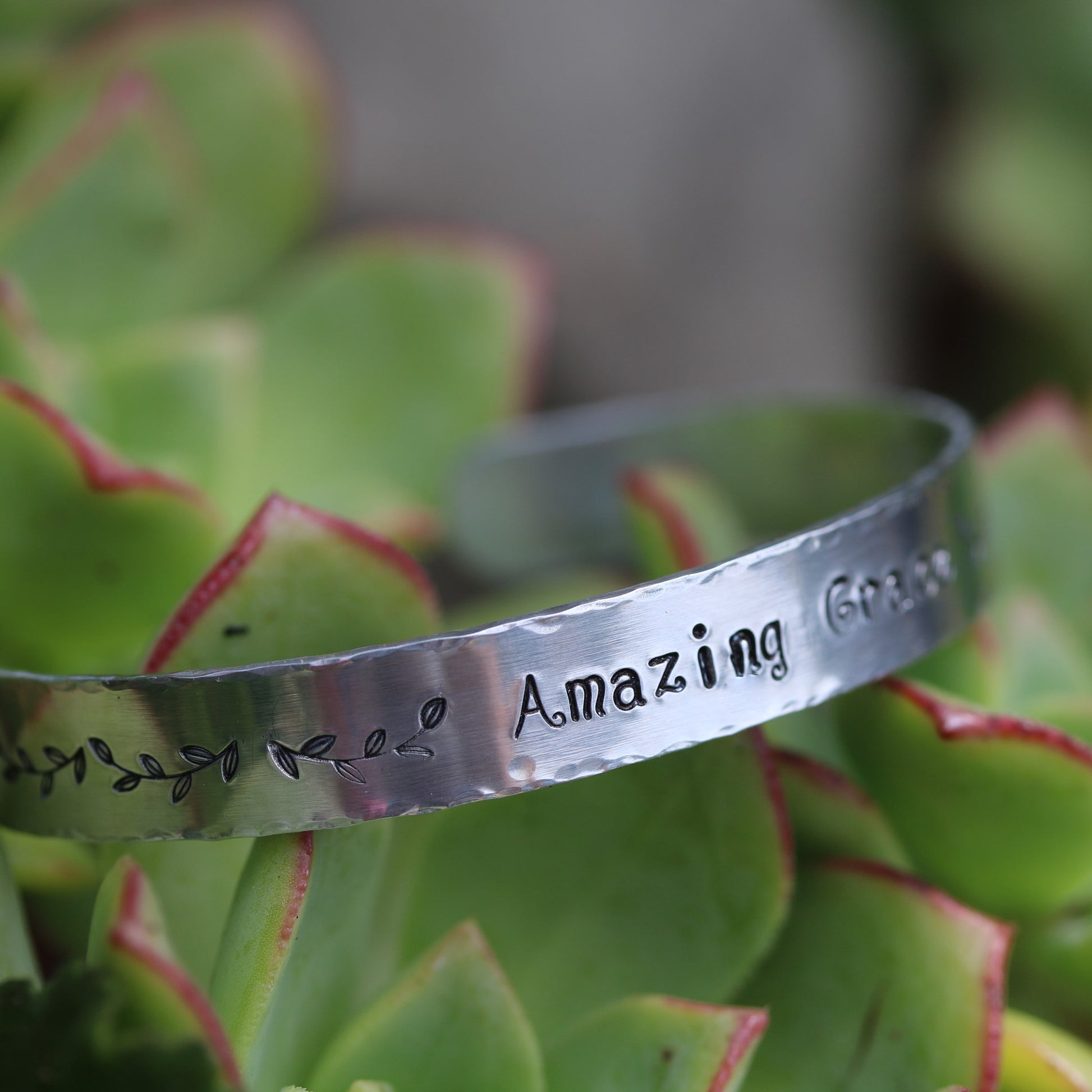 A Sienna Grace Jewelry silver bracelet hand-stamped with ‘Amazing Grace’ sits on a green succulent