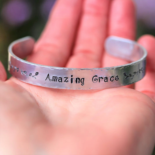 A Sienna Grace Jewelry silver bracelet hand-stamped with ‘Amazing Grace’ sits on a person’s palm